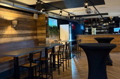 Salle lumineuse avec parquet clair, table et chaises au centre, escalier blanc, machine à coudre ancienne et tableau au mur.