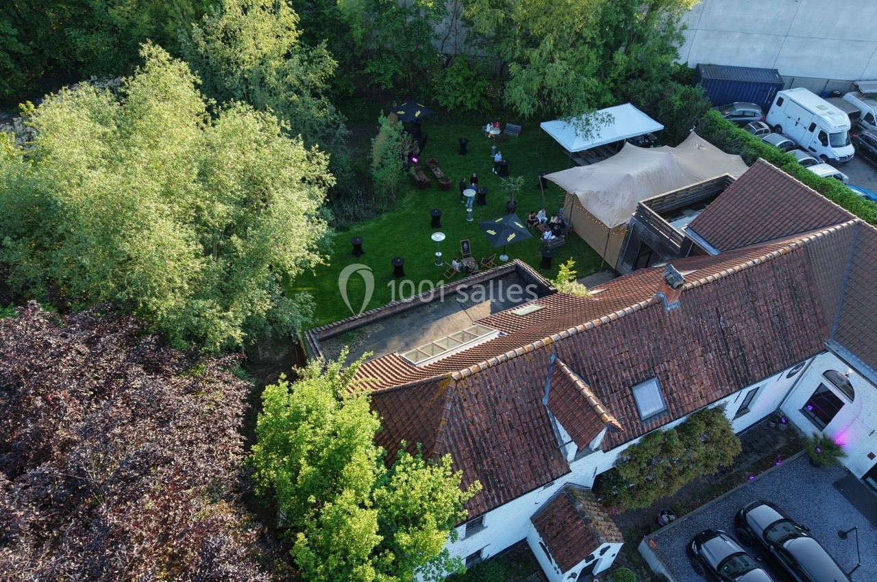 Vue aérienne d'une maison avec un jardin aménagé, des tables, des arbres et des personnes réunies en extérieur.