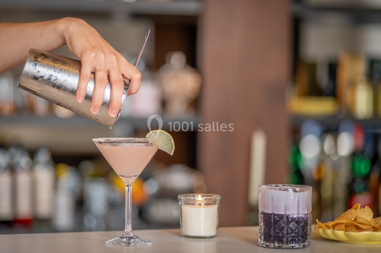 Une main verse un cocktail rose dans un verre décoré d'une tranche de citron vert, avec des boissons et snacks en arrière…