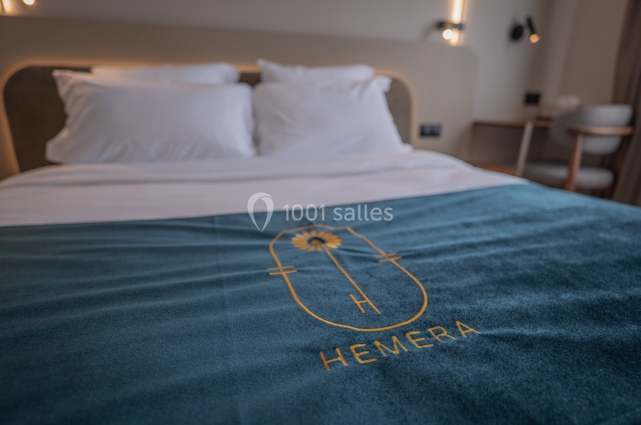 Lit double avec draps blancs et couverture bleue brodée du logo ’Hemera’, dans une chambre moderne et lumineuse.