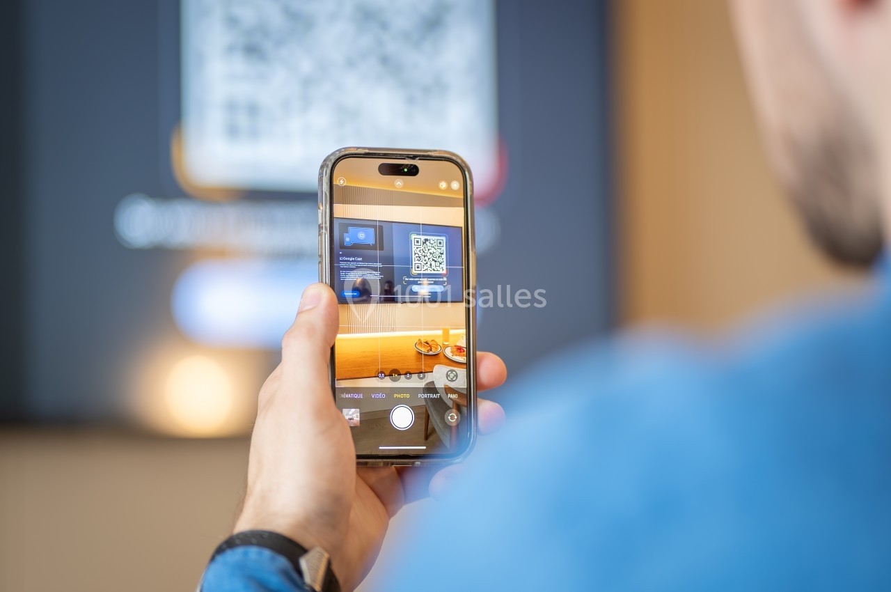 Un homme scanne un code QR affiché sur un écran à l'aide de son smartphone dans un environnement intérieur.