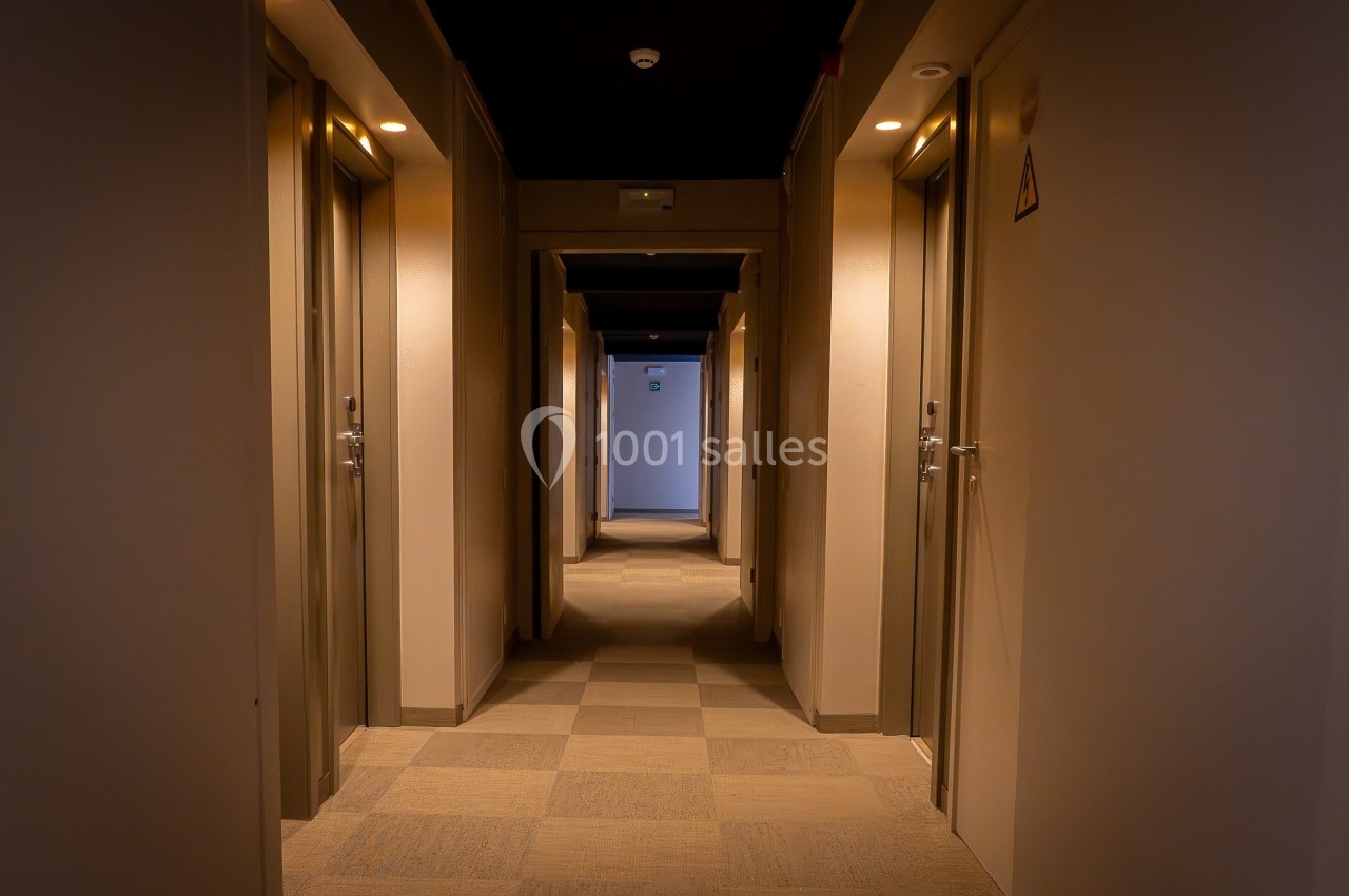 Couloir d'hôtel éclairé avec plusieurs portes de chambres alignées de chaque côté et un sol en carrelage.