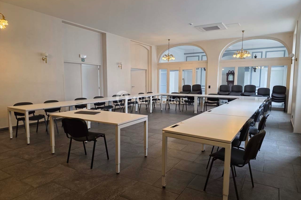 Salle de réunion lumineuse avec plusieurs tables blanches disposées en U et des chaises noires.