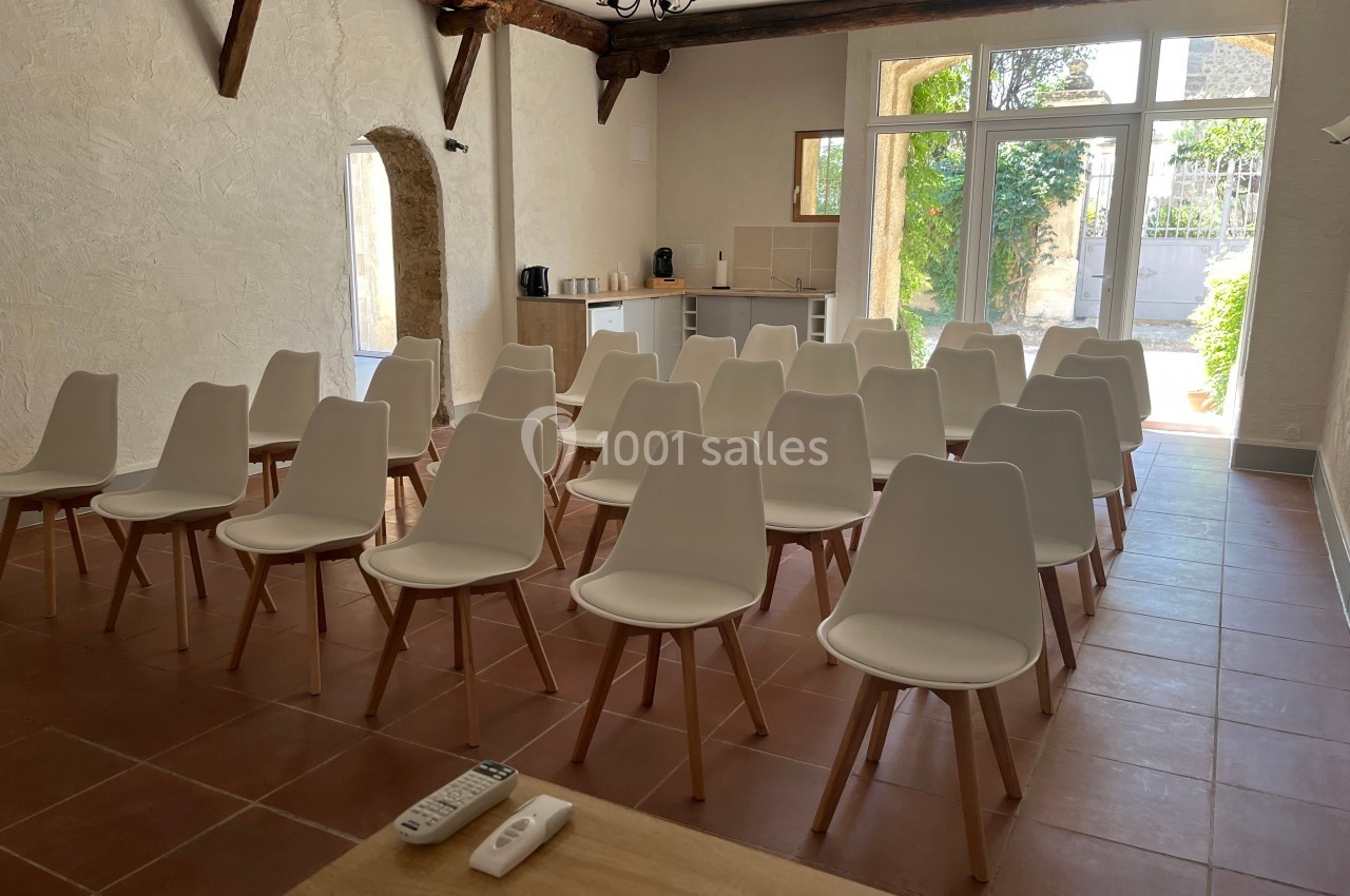 Salle lumineuse avec des chaises blanches alignées, un coin cuisine en arrière-plan et une grande baie vitrée donnant sur un…