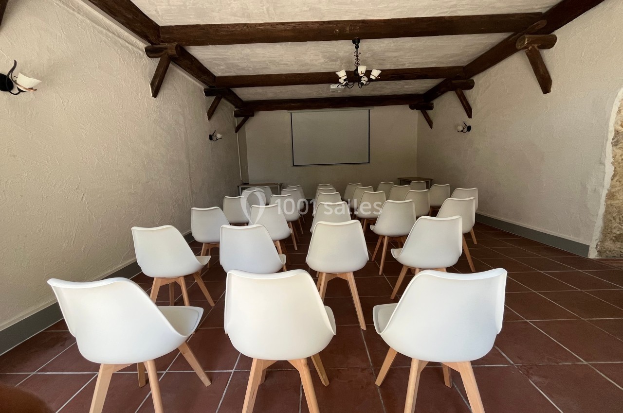 Salle de réunion avec des chaises blanches alignées face à un écran de projection, poutres apparentes au plafond.