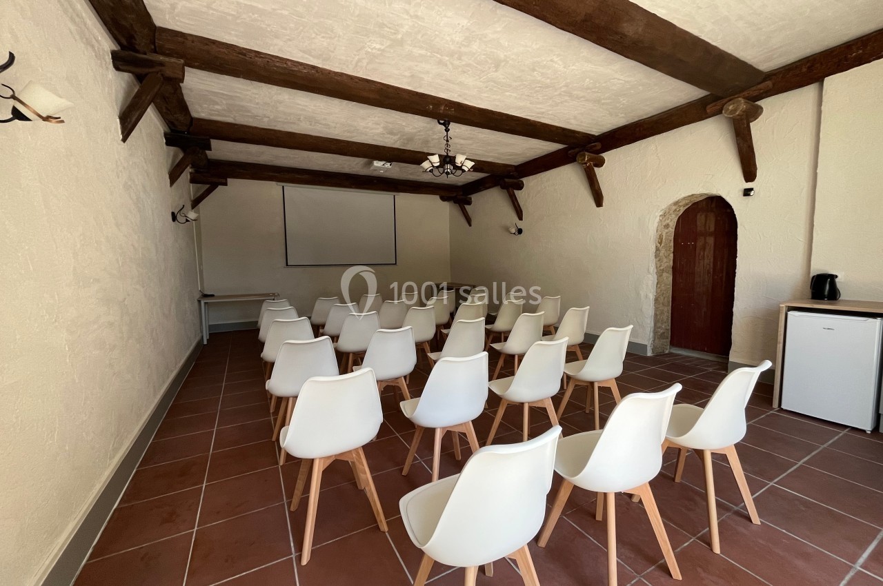 Salle de réunion avec des chaises blanches alignées face à un écran de projection, dans un espace aux poutres apparentes.