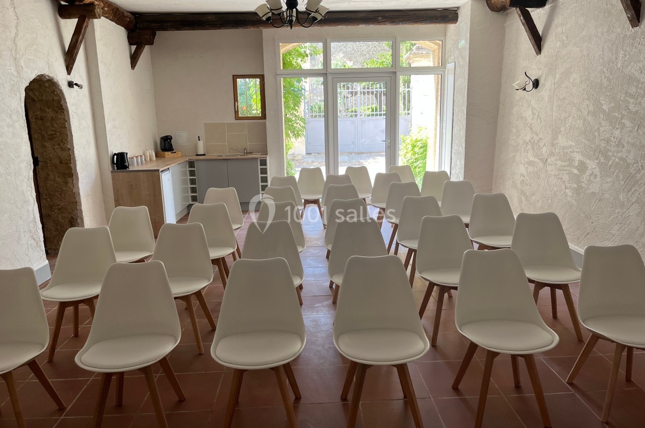 Salle lumineuse avec des chaises blanches alignées face à une petite cuisine et une baie vitrée donnant sur un jardin.