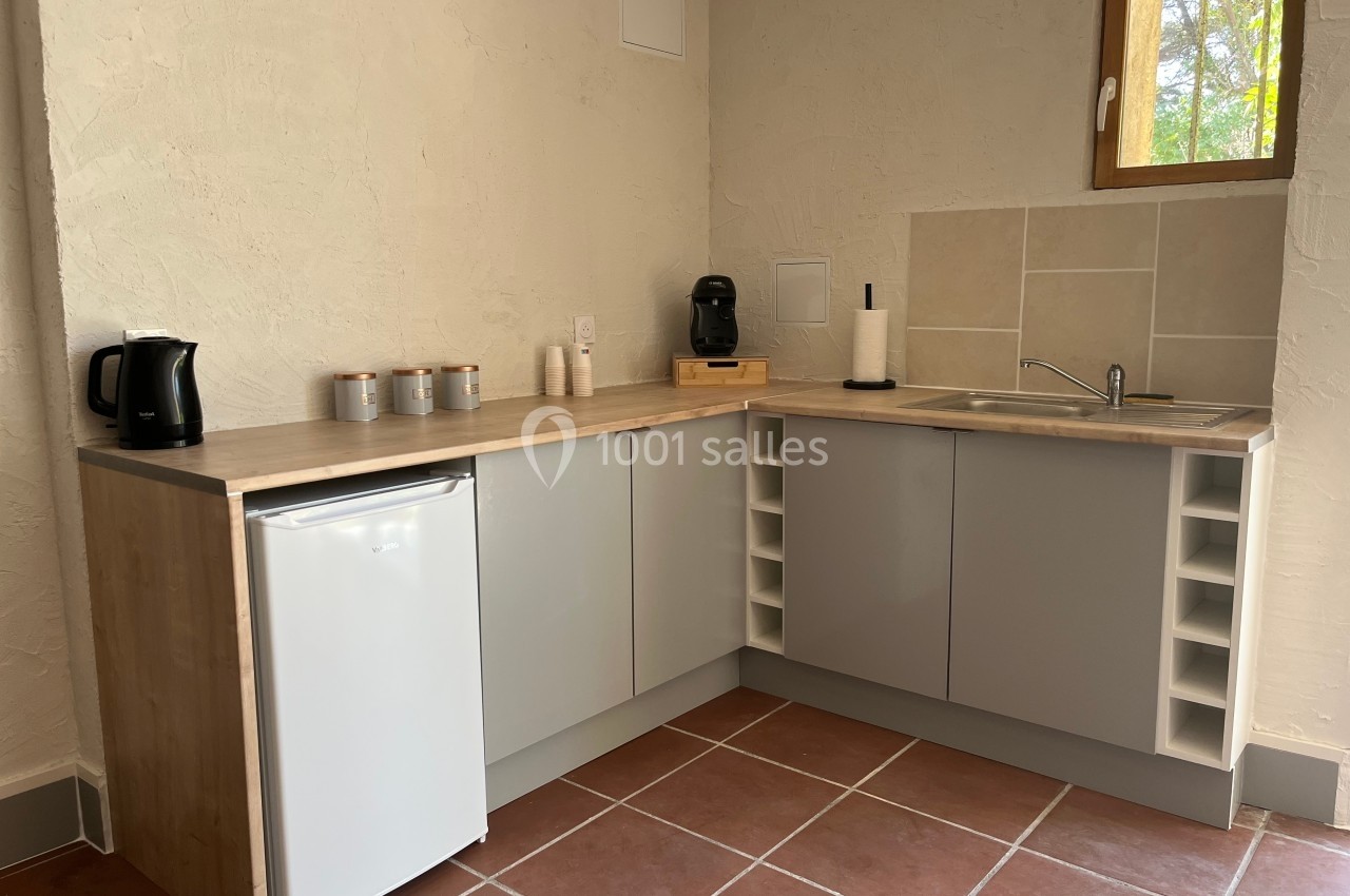 Cuisine compacte avec plan de travail en bois, évier, réfrigérateur, cafetière, et rangements intégrés sur carrelage rouge.