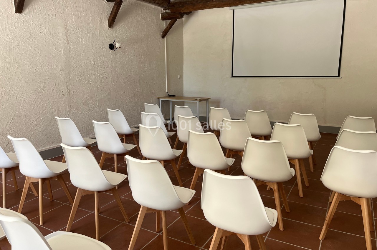 Salle de réunion avec des chaises blanches alignées face à un écran de projection sur un mur clair.