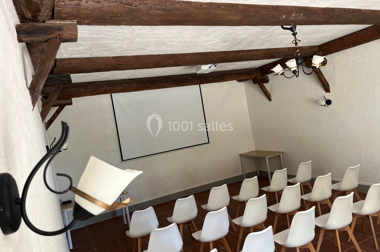 Salle équipée de chaises blanches, d'un projecteur et d'un écran, avec poutres apparentes au plafond.