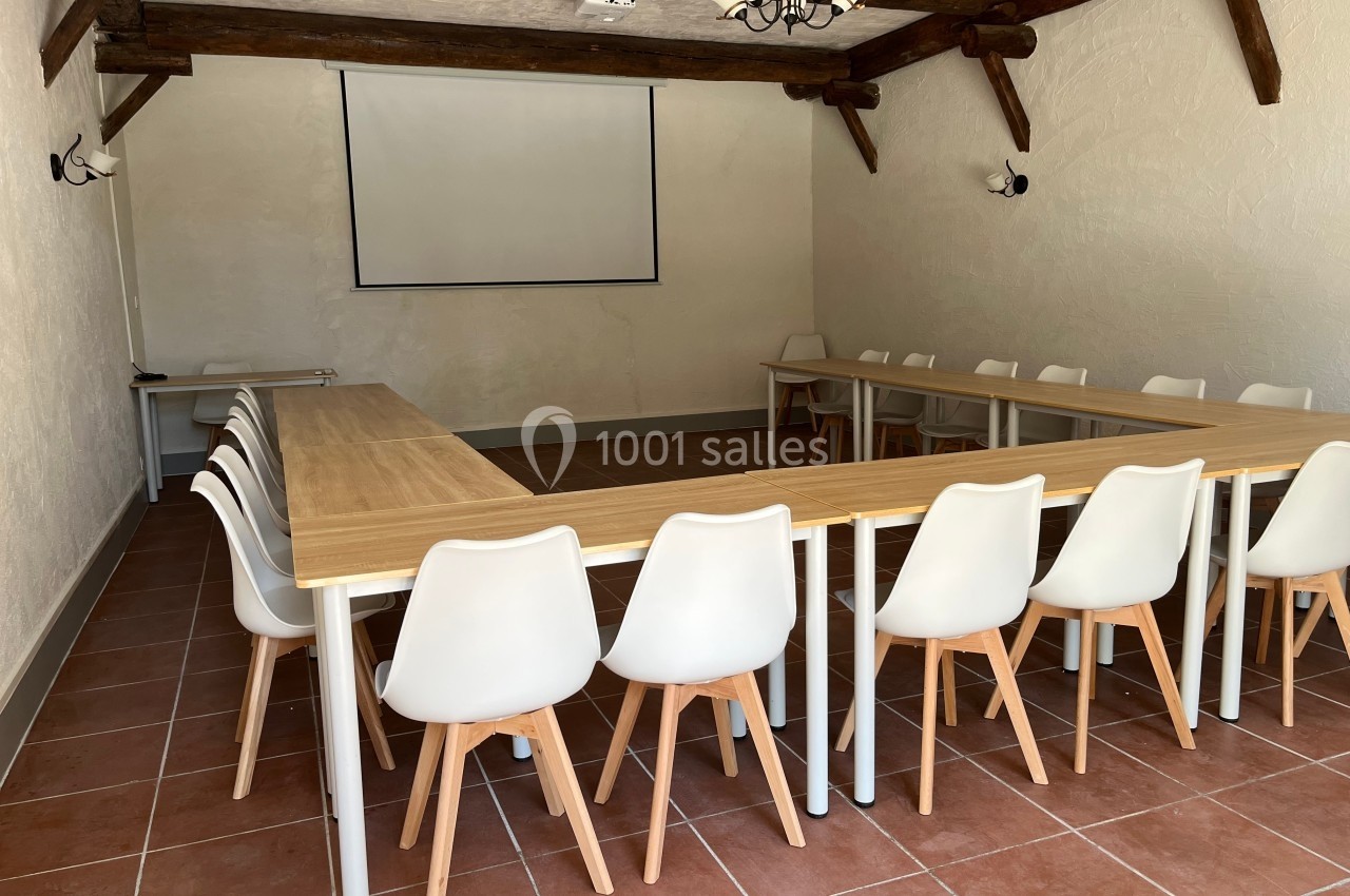 Salle de réunion avec tables disposées en U, chaises blanches, écran de projection et poutres apparentes au plafond.