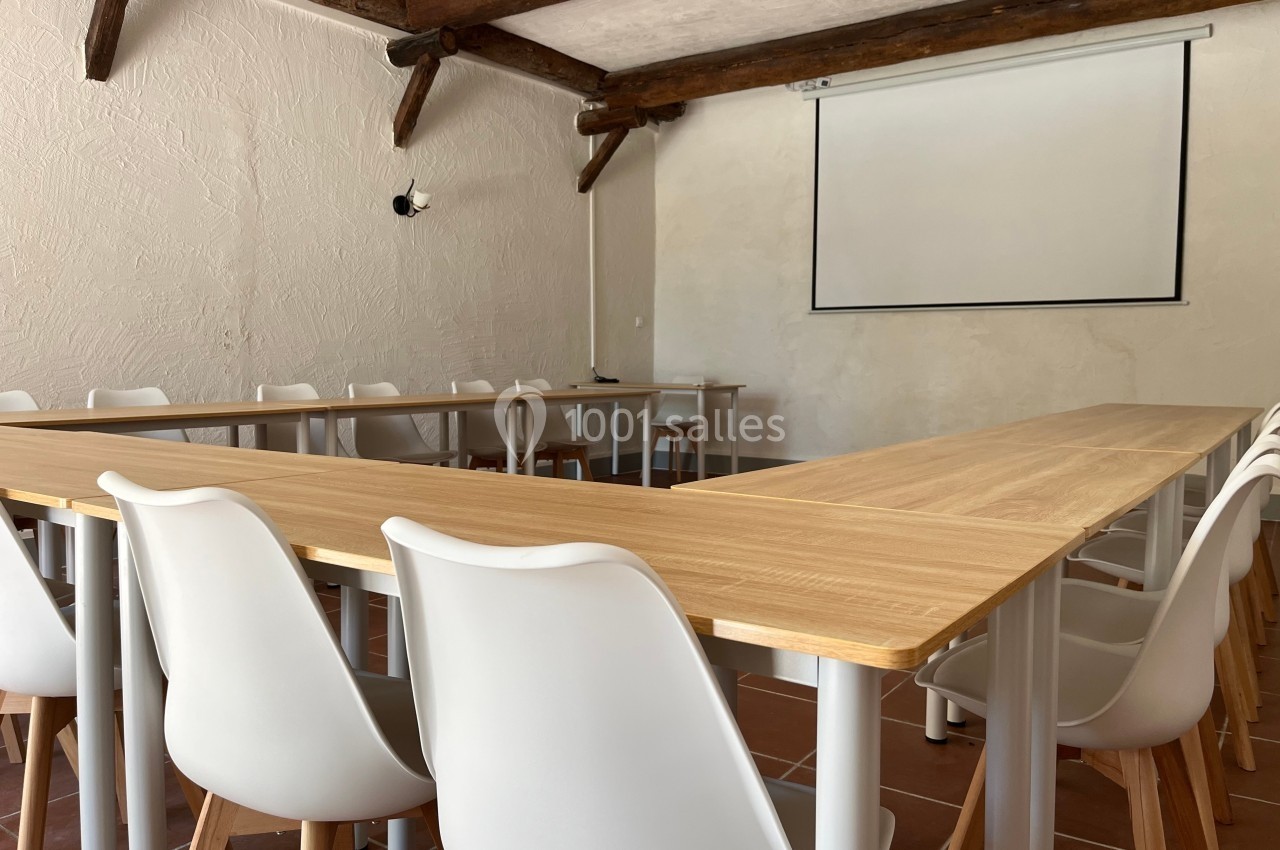 Salle de réunion lumineuse avec tables en bois disposées en U, chaises blanches et écran de projection mural.