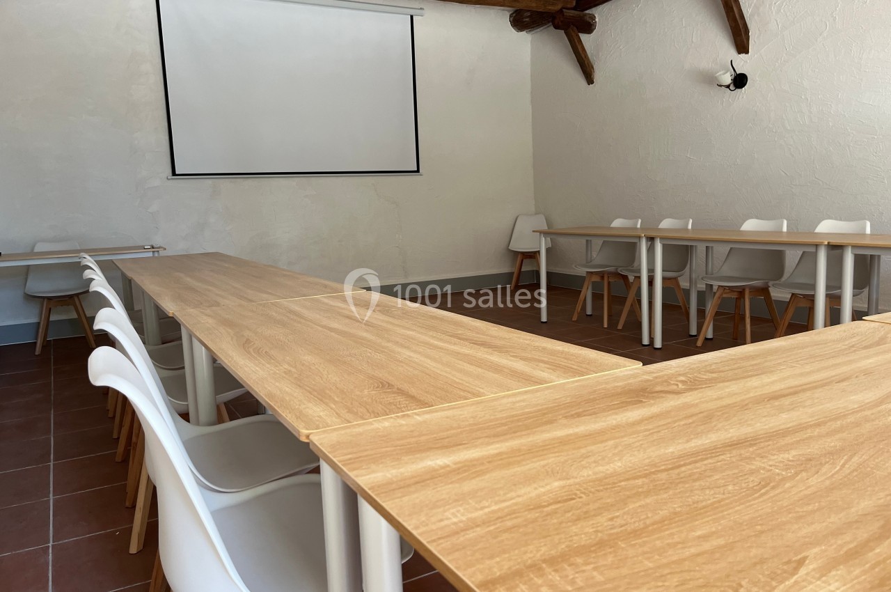 Salle de réunion avec tables en bois clair, chaises blanches et écran de projection fixé au mur.