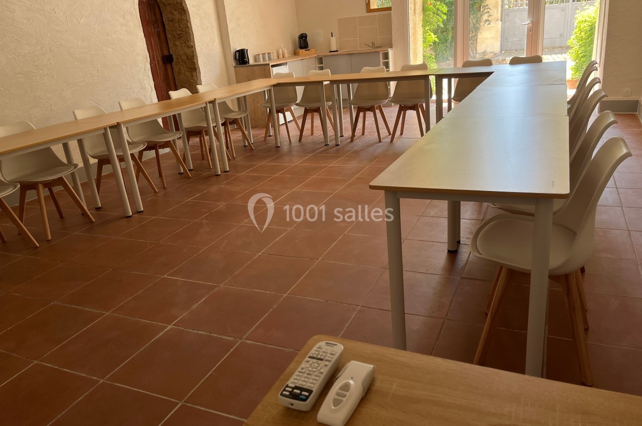 Salle lumineuse avec tables et chaises disposées en U, sol en carrelage, porte en bois et baie vitrée donnant sur un jardin.