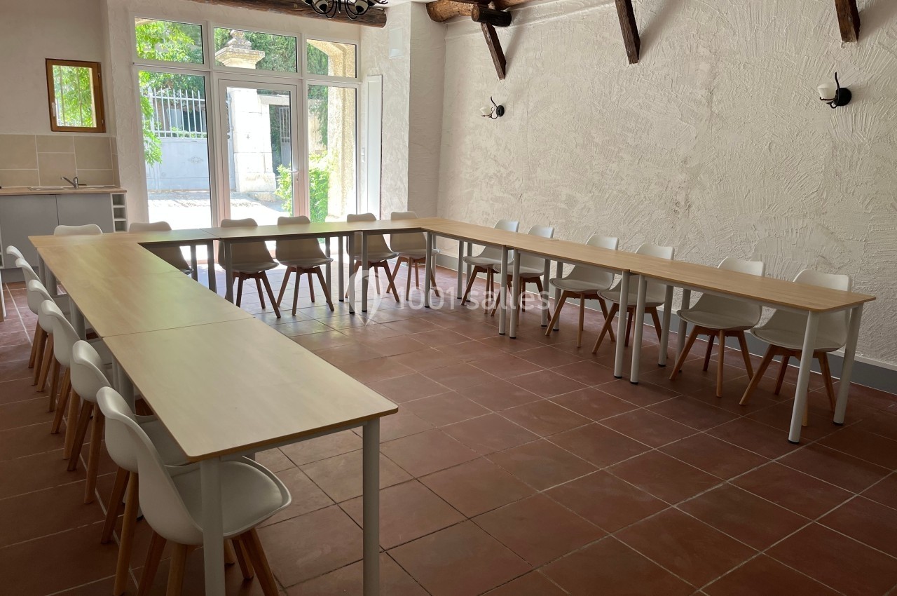 Salle lumineuse avec tables en U, chaises blanches, carrelage au sol, murs clairs et poutres apparentes.