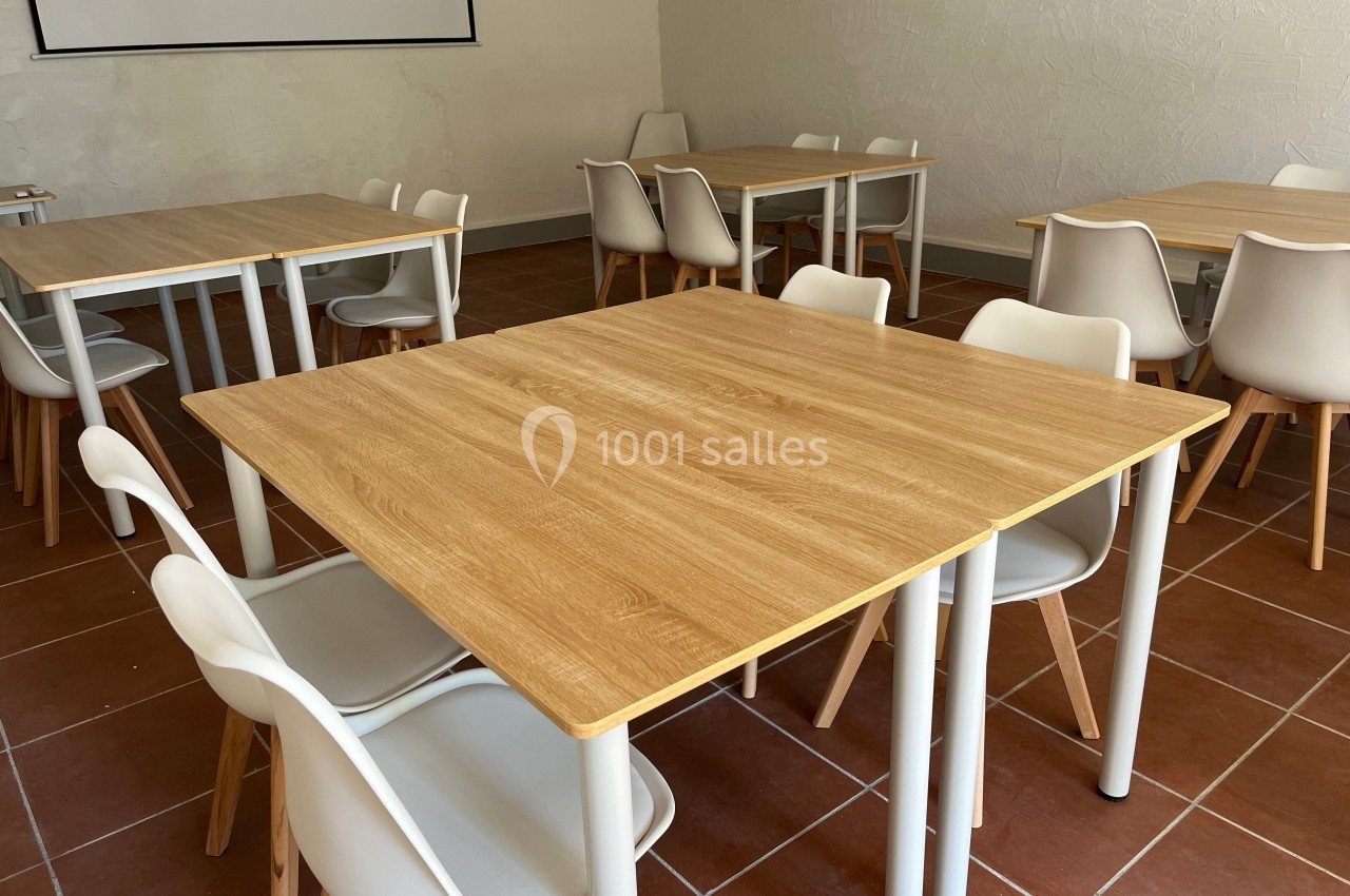 Salle avec des tables en bois clair et des chaises blanches, disposées sur un sol en carrelage brun.