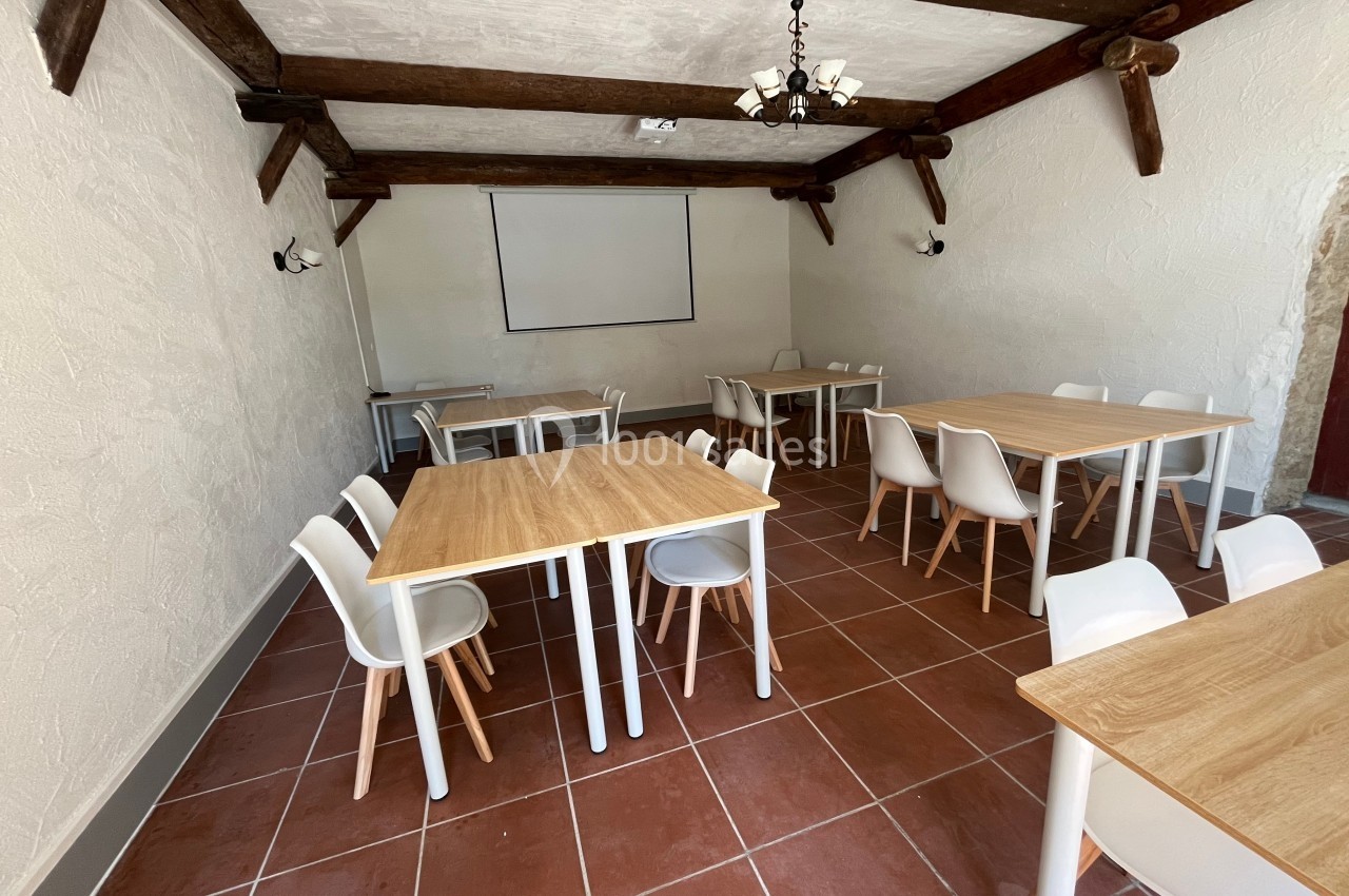 Salle lumineuse avec tables en bois clair, chaises blanches et écran de projection sur un mur blanc.