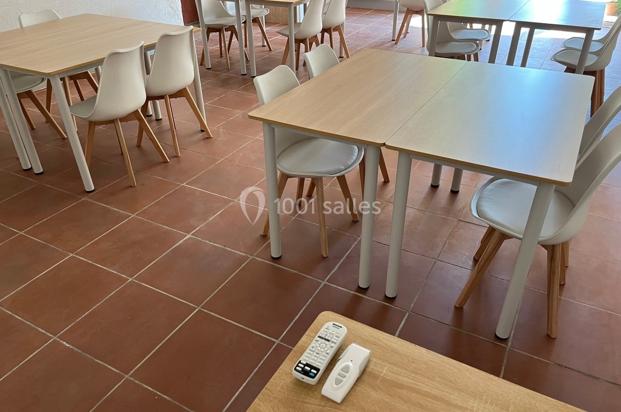 Salle avec des tables en bois clair, chaises blanches et un sol en carrelage rouge, télécommandes sur une table.