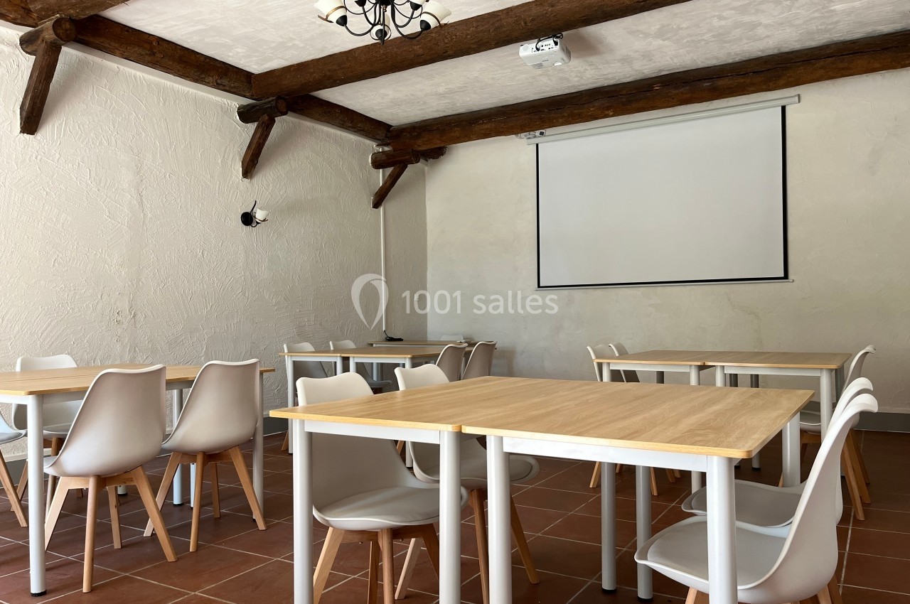 Salle lumineuse avec tables en bois clair, chaises blanches, poutres apparentes et écran de projection mural.