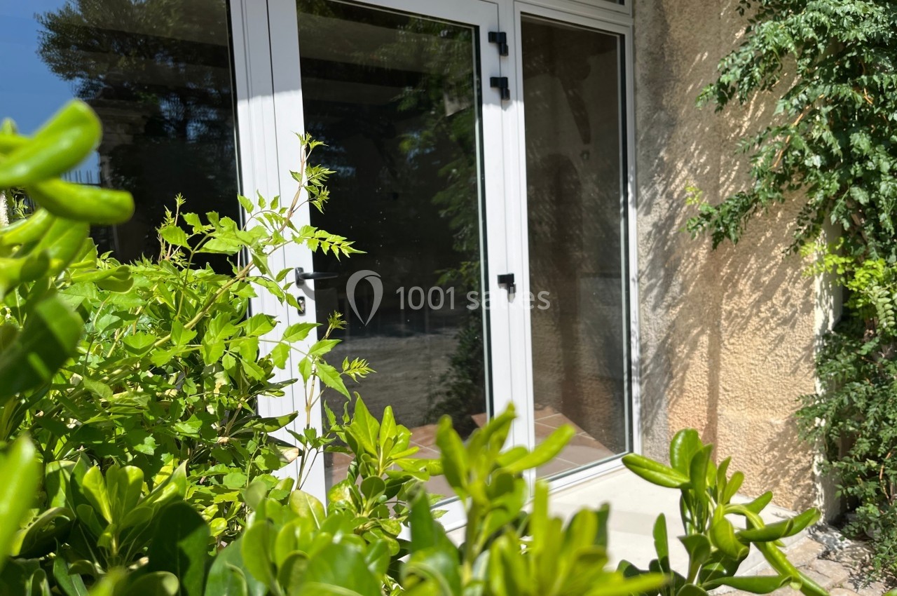 Porte-fenêtre blanche donnant sur un extérieur ensoleillé, entourée de végétation et d'un mur en pierre.