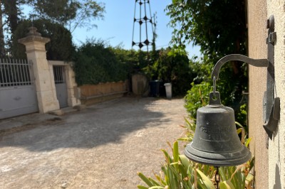 Cloche en métal fixée à un mur, avec une allée gravillonnée, des plantes et un portail en arrière-plan.
