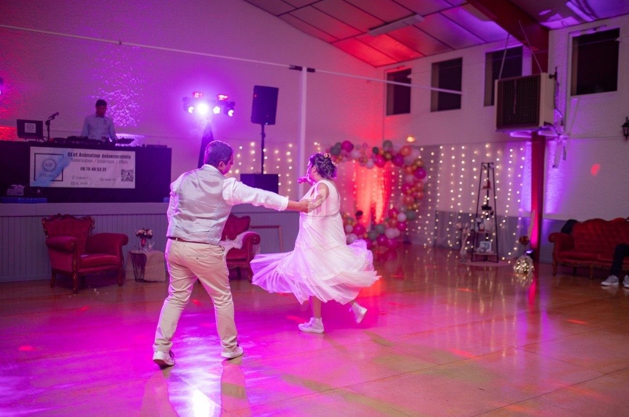 Un couple danse sur une piste éclairée de lumières colorées, avec un DJ en arrière-plan.