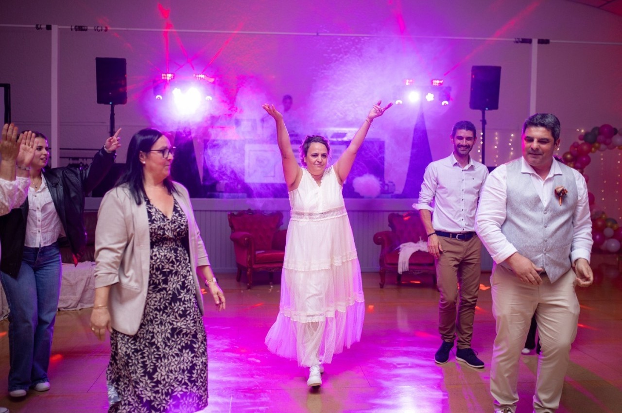Personnes dansant dans une salle éclairée par des lumières colorées, avec une femme en robe blanche au centre levant les…