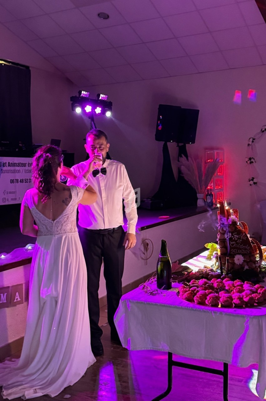 Un couple en tenue de mariage partage un moment devant une table garnie de desserts dans une salle éclairée.