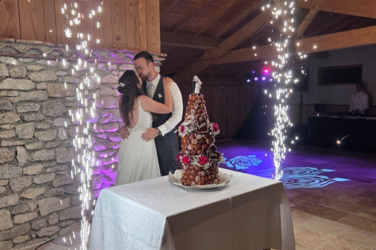 Un couple de mariés s'enlace près d'une pièce montée éclairée par des étincelles dans une salle en bois et pierre.