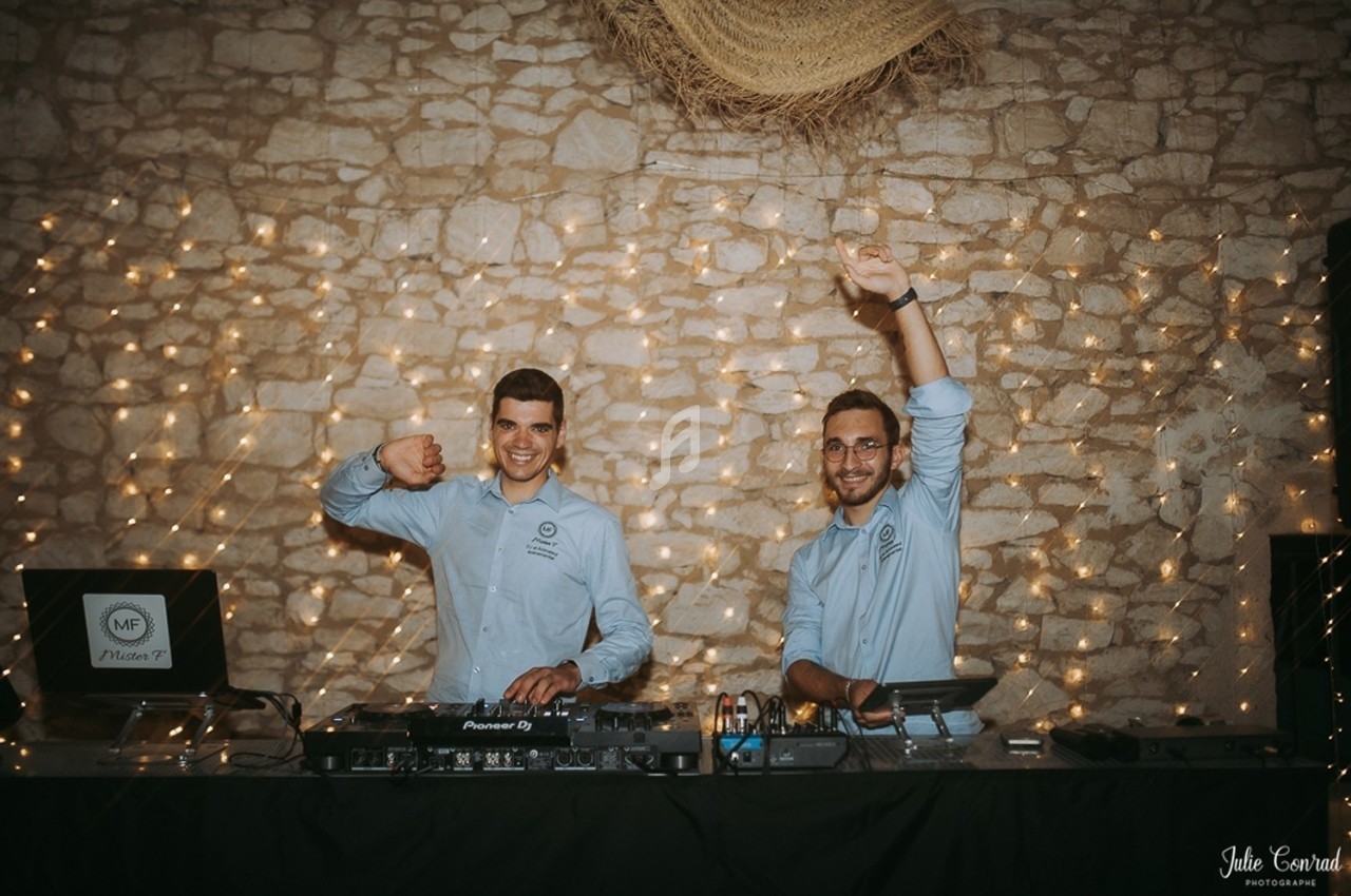 Deux DJ souriants en chemise bleue animent une soirée devant un mur en pierre décoré de guirlandes lumineuses.