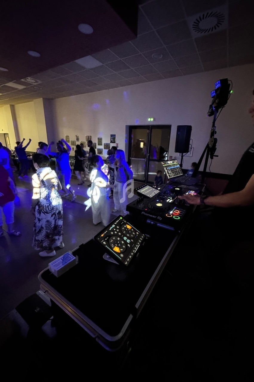Un DJ mixe sur une table de mixage tandis que des personnes dansent dans une salle éclairée par des lumières colorées.