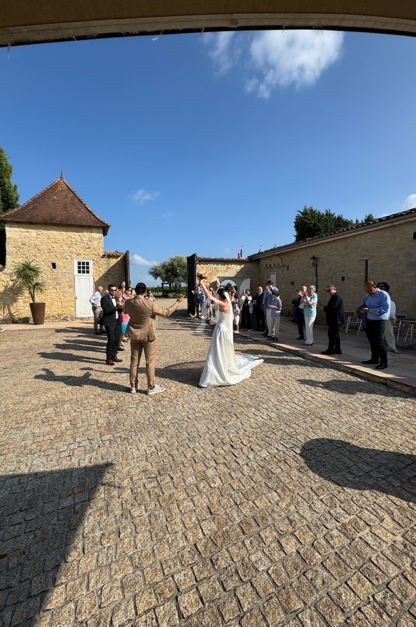 Un couple de mariés danse dans une cour pavée entourée d'invités applaudissant sous un ciel dégagé.
