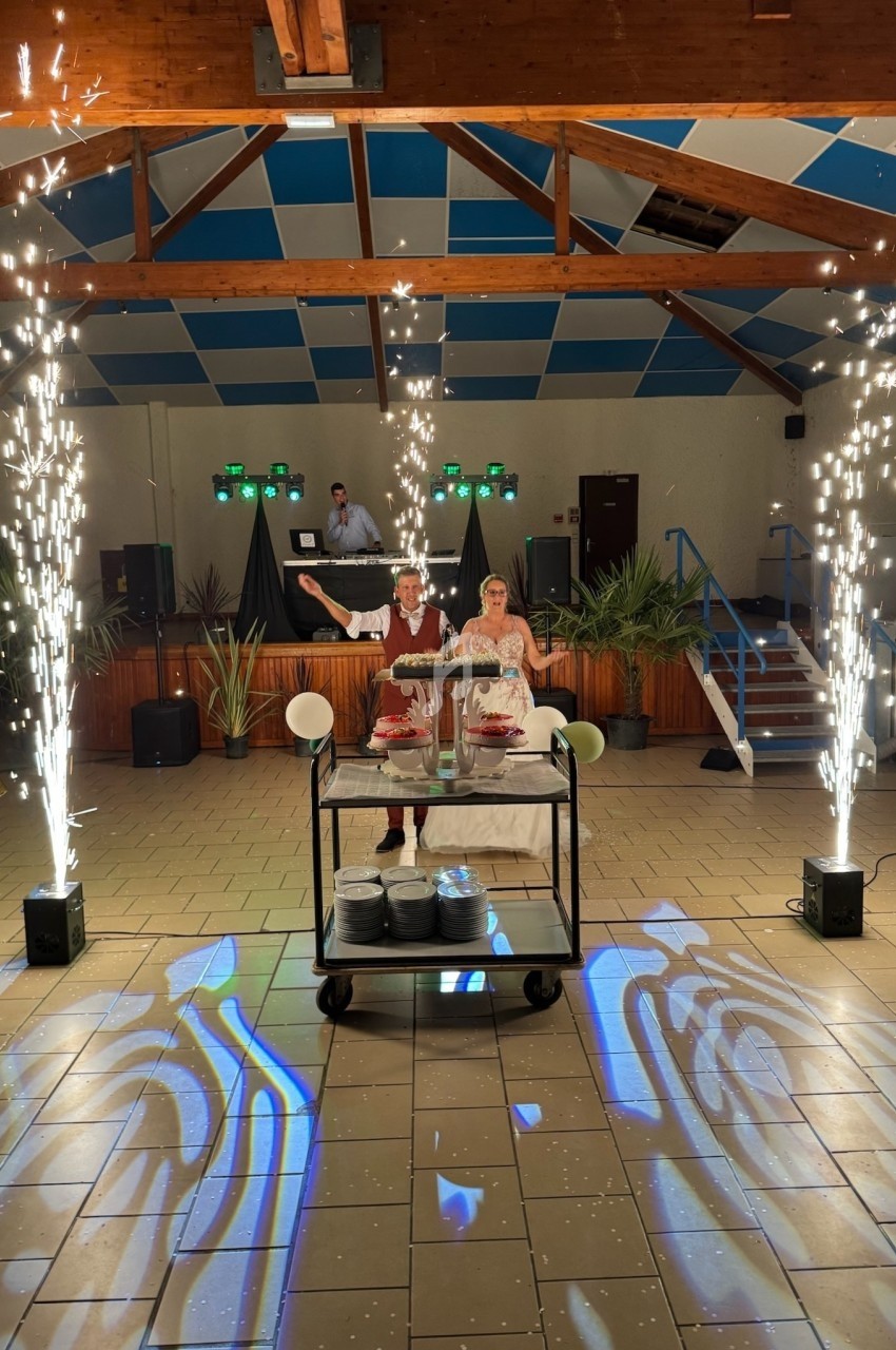 Deux personnes debout derrière un chariot avec des gâteaux, entourées de fontaines d'étincelles dans une salle festive.