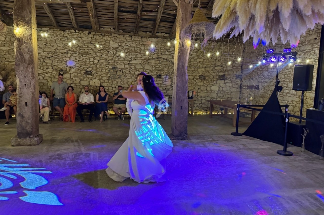 Une mariée danse seule sous des lumières colorées dans une salle en pierre avec des invités assis en arrière-plan.