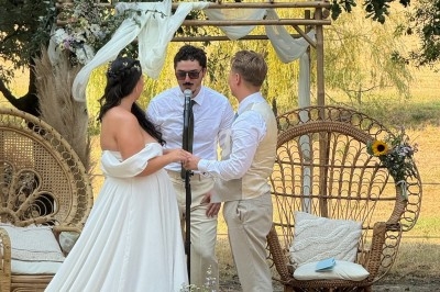 Un couple échange ses vœux devant un officiant, sous une arche décorée de fleurs, dans un cadre extérieur champêtre.
