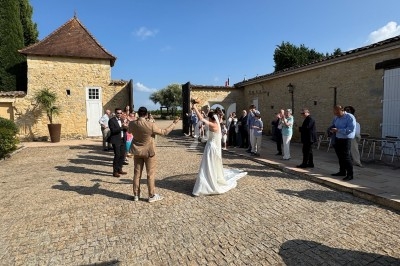 Un couple de mariés danse en extérieur, entouré d'invités applaudissant dans une cour pavée sous un ciel bleu.