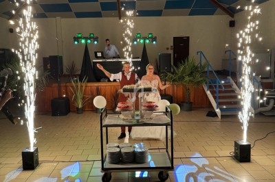 Deux personnes debout derrière un chariot avec des gâteaux, entourées de fontaines d'étincelles dans une salle festive.