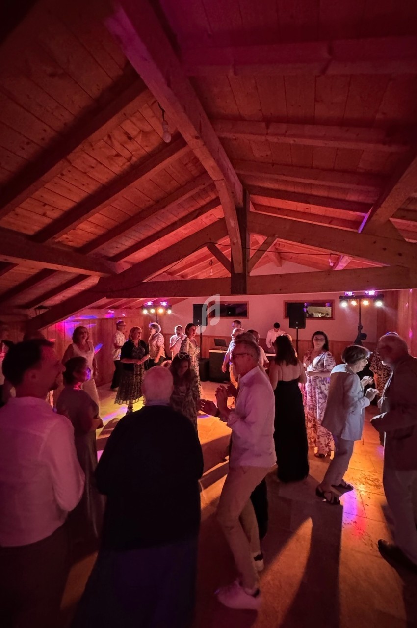 Des personnes dansent dans une salle en bois éclairée par des lumières colorées, sous un plafond avec poutres apparentes.