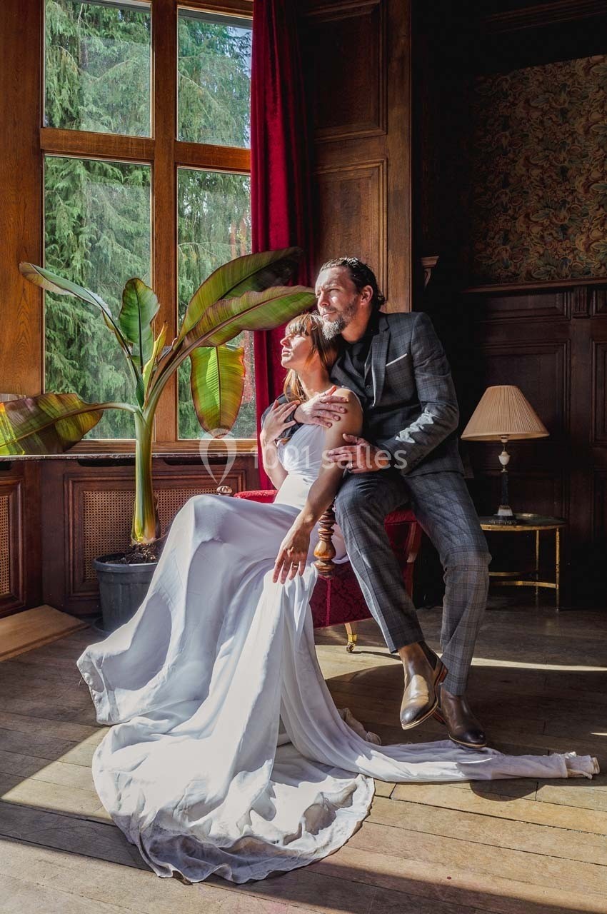 Un couple élégant pose dans une pièce lumineuse avec des boiseries, près d'une grande fenêtre et d'une plante tropicale.