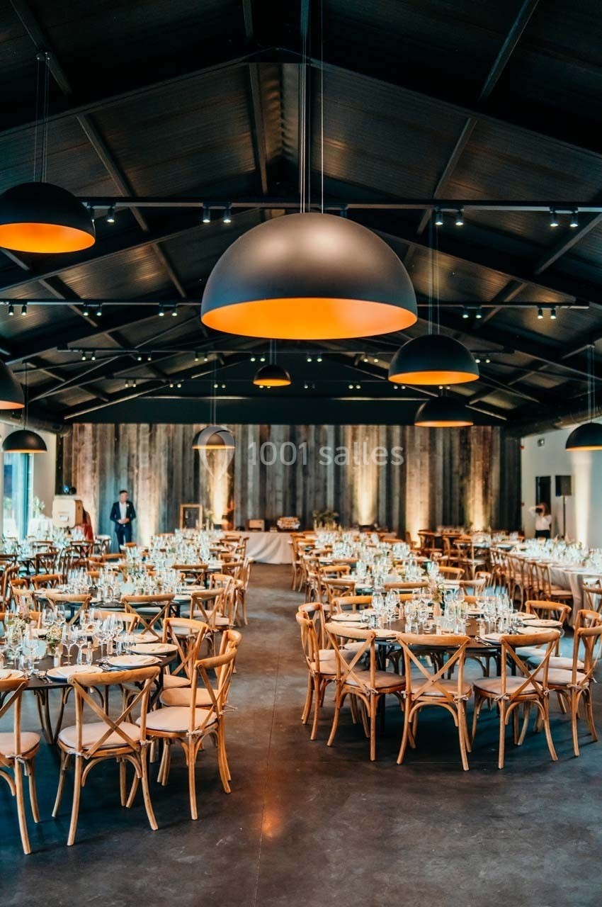 Salle de réception élégante avec tables rondes dressées, chaises en bois et suspensions lumineuses modernes.