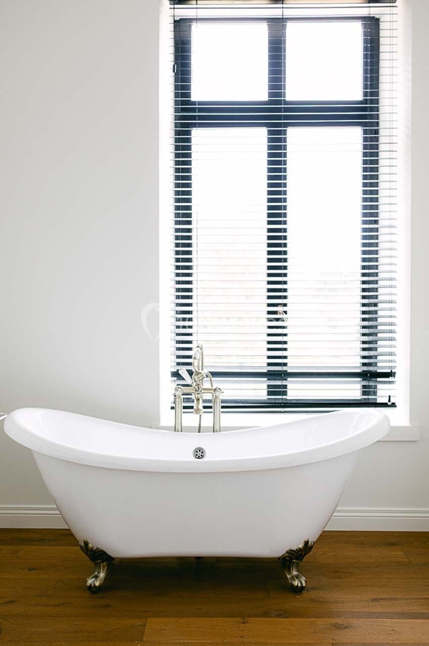 Baignoire blanche sur pieds devant une fenêtre avec stores vénitiens, posée sur un parquet en bois clair.