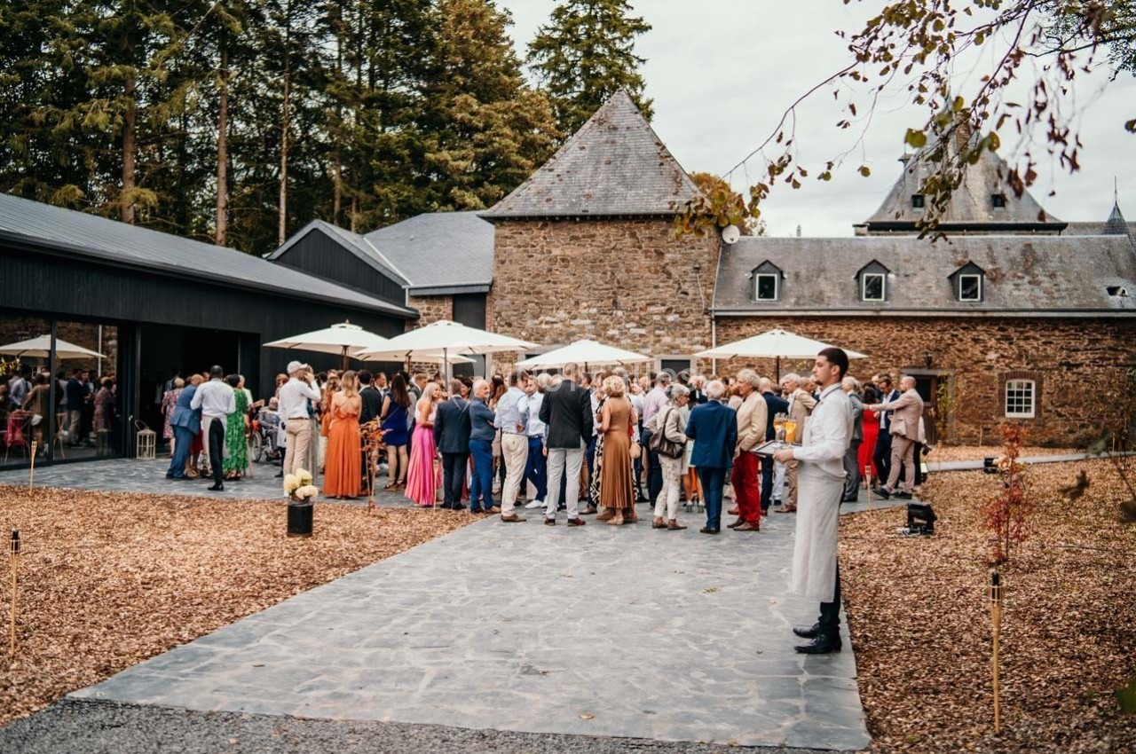 Groupe de personnes rassemblées en extérieur devant un bâtiment en pierre avec parasols et arbres en arrière-plan.