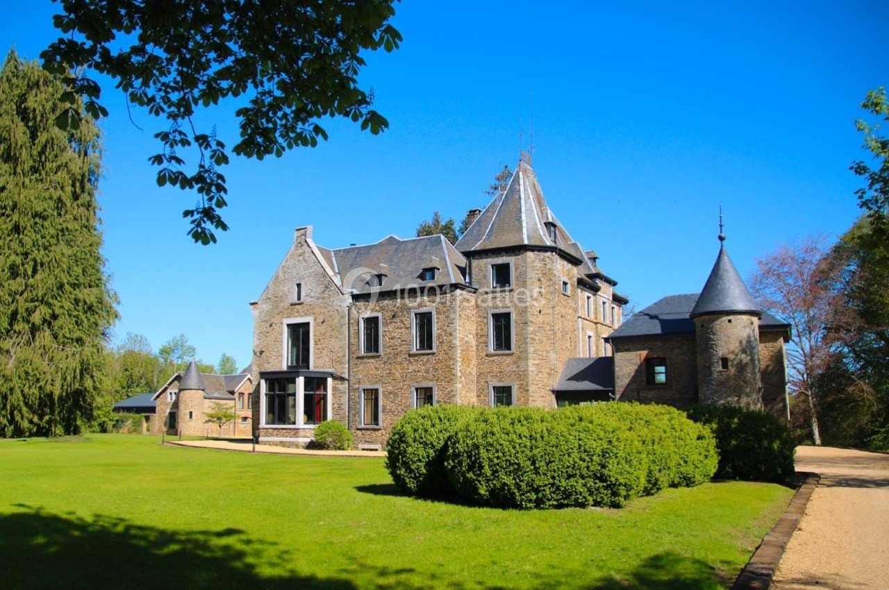Château en pierre entouré de pelouses et d'arbres, sous un ciel bleu clair.
