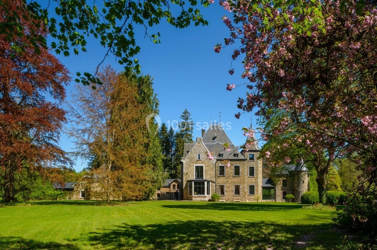 Manoir en pierre entouré d'arbres colorés et d'une pelouse verdoyante sous un ciel bleu clair.