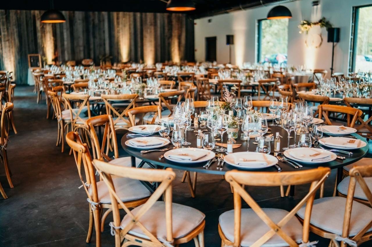 Salle de réception décorée avec des tables rondes dressées, chaises en bois et lumière naturelle entrant par les fenêtres.