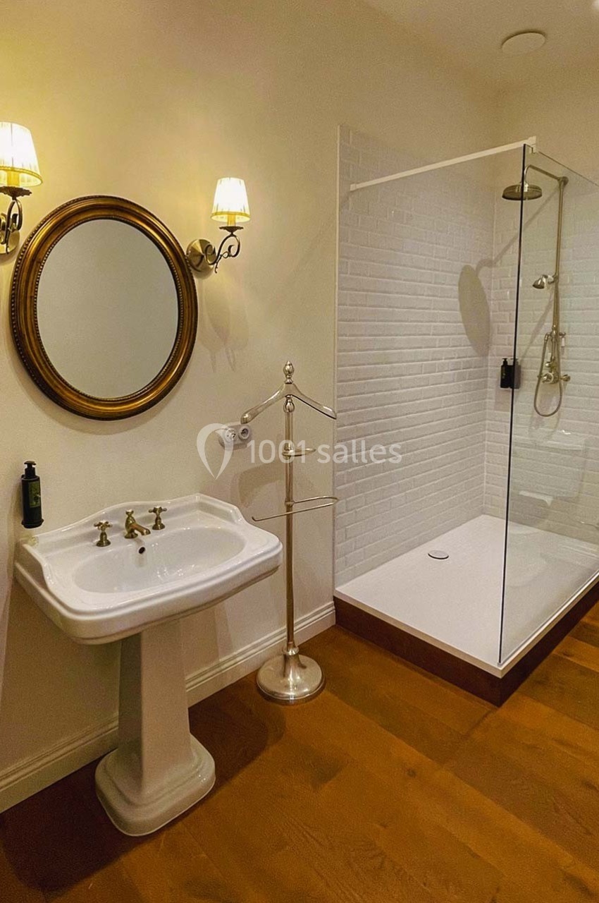 Salle de bain avec lavabo sur pied, miroir rond, douche à l'italienne vitrée et sol en parquet.