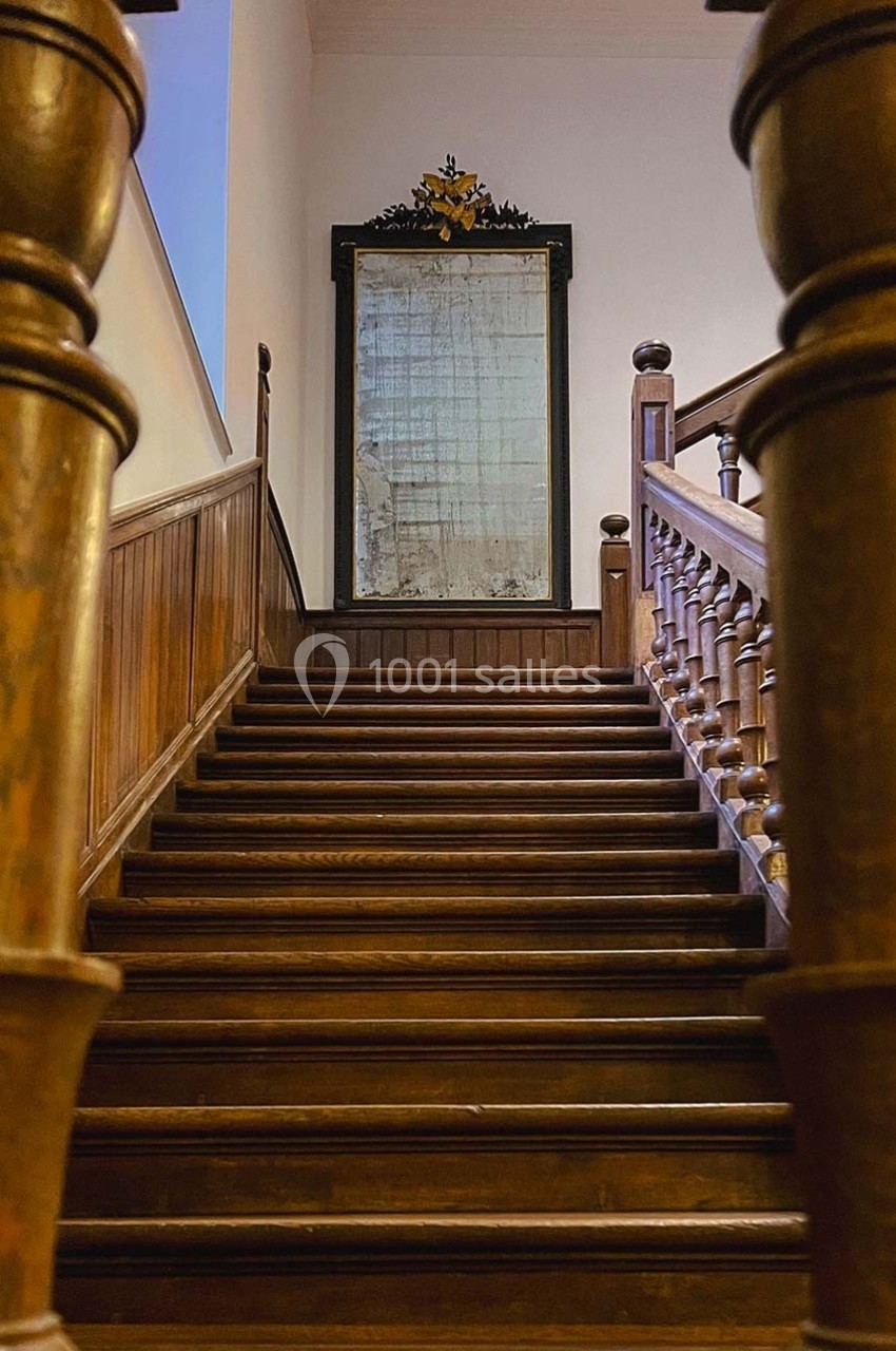Escalier en bois ancien avec une rampe sculptée, menant à un grand miroir encadré sur un mur clair.