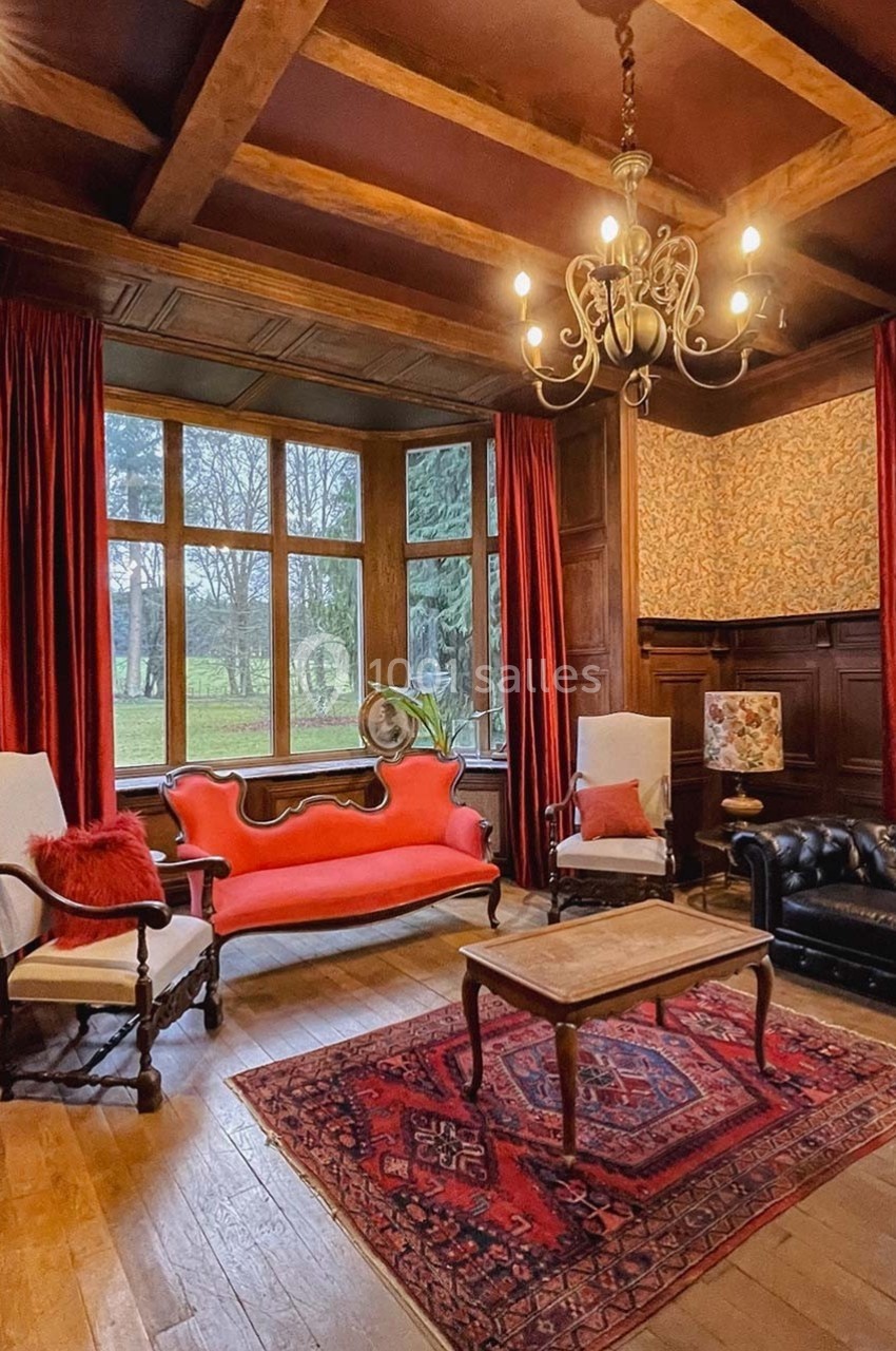 Salon chaleureux avec canapé rouge, fauteuils, tapis persan, lustre et grandes fenêtres donnant sur un jardin.