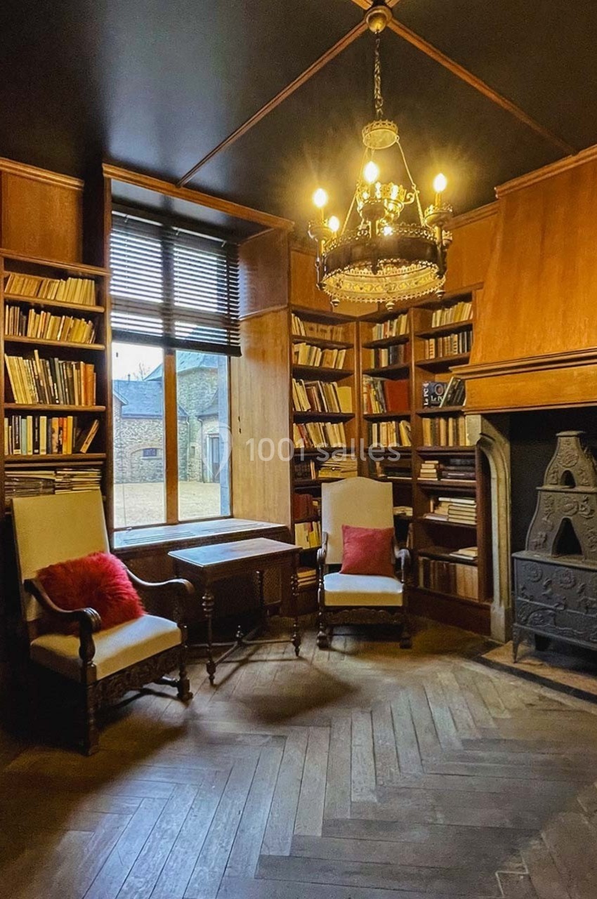 Bibliothèque ancienne avec étagères en bois, fauteuils, cheminée en fonte et lustre, éclairée par une fenêtre.