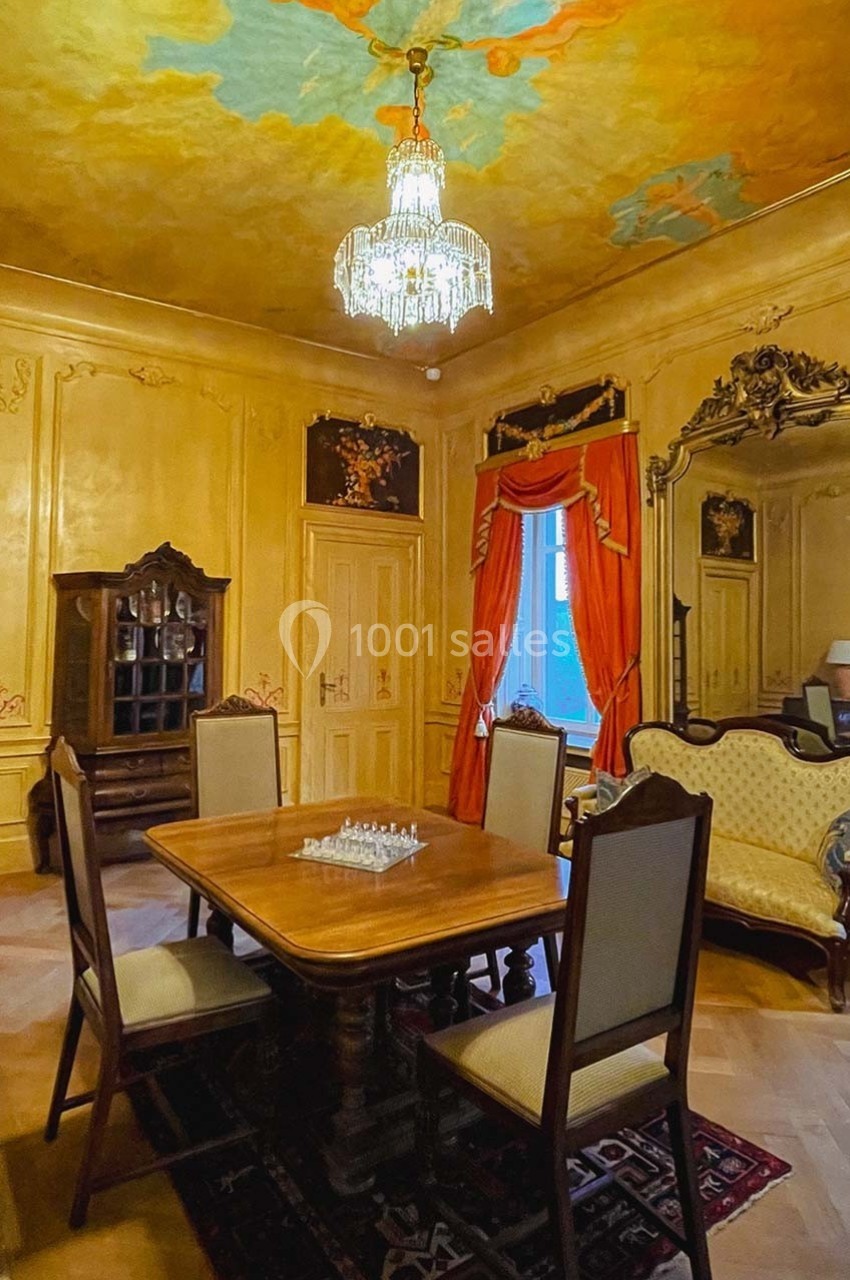 Salle à manger ancienne avec mobilier en bois, lustre en cristal, fresque au plafond et rideaux rouges encadrant une fenêtre.